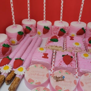 May include: A collection of pink and white treats, including marshmallow pops with strawberry and daisy decorations, pink-frosted rice crispy treats with a red "1", and pink-covered pretzel rods. Round cookies with strawberry designs and text are also visible.