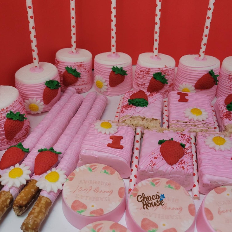 May include: A collection of pink and white treats, including marshmallow pops with strawberry and daisy decorations, pink-frosted rice crispy treats with a red "1", and pink-covered pretzel rods. Round cookies with strawberry designs and text are also visible.