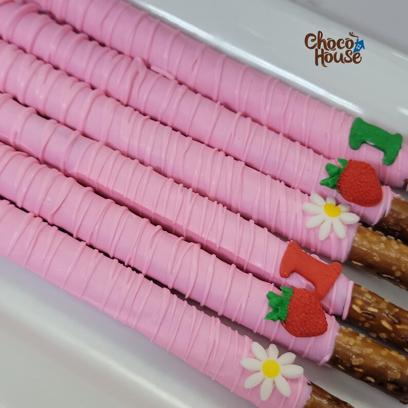 May include: Pink chocolate-covered pretzel rods with white drizzle. Decorated with strawberry and daisy-shaped candies. The image includes the text "Choco House" in the upper right corner. The pretzels are arranged on a white tray.
