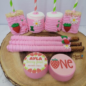 May include: A collection of pink-frosted treats, including Rice Krispie treats, marshmallow pops with straws, and chocolate-covered pretzel rods. Decorated with strawberries and daisies, and two round treats with text that reads "AYLA berry first BIRTHDAY PARTY" and "ONE".