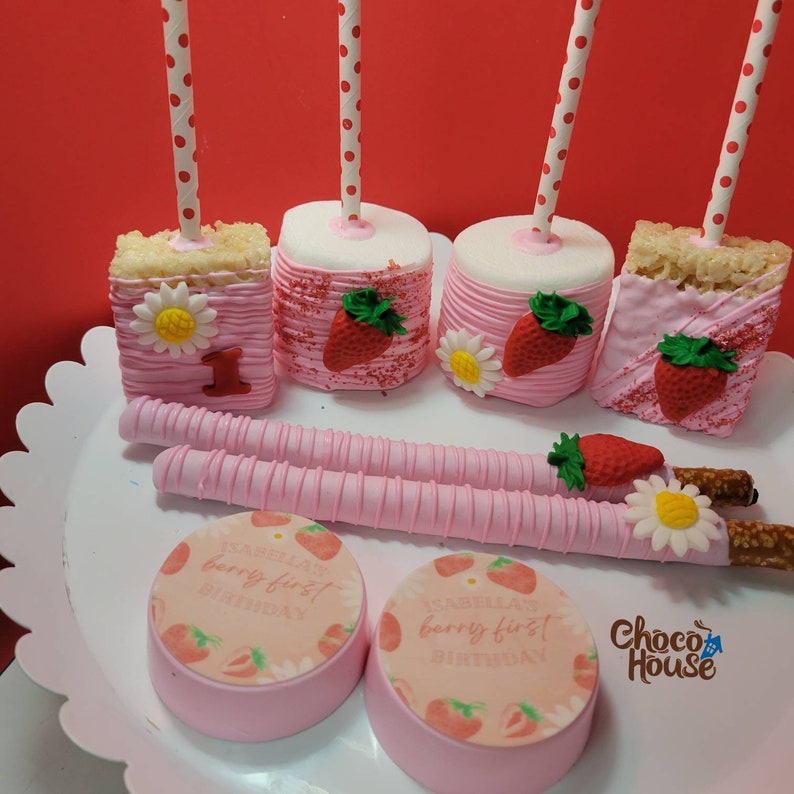 May include: A collection of pink and white treats, including marshmallow pops, rice crispy treats, and chocolate-covered cookies. The treats are decorated with strawberries, daisies, and the text "Isabella's berry first birthday". The treats are on a white scalloped tray.