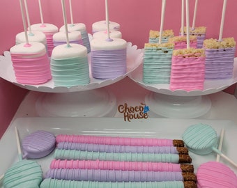 May include: Pastel-colored treats on display. Marshmallow pops in pink, blue, and purple, and rice crispy treats in similar hues. Chocolate-covered pretzels and lollipops complete the sweet assortment, all arranged on white serving platters.