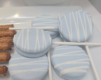 May include: A close-up of blue and white candy-coated Oreo cookies on sticks, presented on a white plate. The cookies are adorned with white stripes. Pretzels are visible on the left side of the plate.