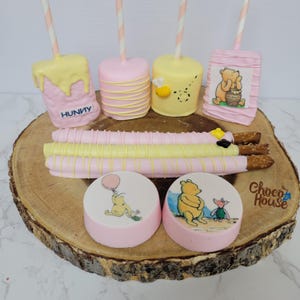 May include: A collection of sweets including four cake pops on striped sticks, three chocolate-covered pretzel rods, and two round cookies. The treats feature pink and yellow icing, honey pot imagery, and illustrations of a bear and a piglet.