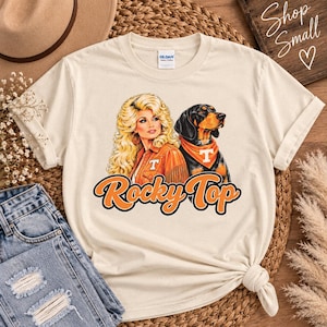 May include: A cream-colored short-sleeve t-shirt featuring a graphic print of a blonde woman with voluminous hair wearing an orange jacket and a black and tan hound dog wearing an orange bandana. The text Rocky Top is printed in orange below.