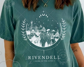 May include: A teal blue t-shirt with a white graphic design of a city with a crescent moon and stars. The text "Rivendell - Home of the Elves" is printed below the graphic. The shirt is being worn by a person.