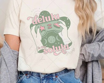May include: A white t-shirt with a green and pink graphic of a video game controller with the text "Delulu is the solutiu".