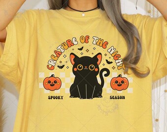 May include: Mustard-colored t-shirt featuring a graphic of a black cat, pumpkins, and the text "CREATURE OF THE NIGHT" and "SPOOKY SEASON". The shirt has short sleeves and a relaxed fit, perfect for Halloween.