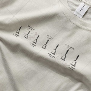 Puede incluir: Camiseta blanca con un gráfico negro de campanas y nombres, incluyendo "Kainna", "Morsakel", "Kirsethi", "Dytrim", "Peloker", "Saranneth" y "Astarrael".