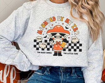 May include: Ash gray sweatshirt with a graphic featuring a mushroom-headed character holding a book, with ghosts and the text "HAUNTED BY MY TBR." Below, it reads "TOO MANY BOOKS TOO LITTLE TIME." The design has a retro, Halloween theme.