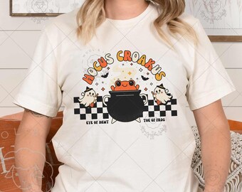 May include: Cream-colored t-shirt featuring a Halloween-themed graphic. The design includes a cauldron with a frog, ghosts, and the text "Hocus Croakus". The shirt has short sleeves and a crew neck.