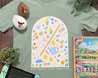 May include: Sage green t-shirt with a graphic of a hoe, watering can, and other farm-related items in a window shape. The text "All About That Hoe Life" is printed below the graphic. The t-shirt is laying on a wooden surface with a Nintendo Switch, a book, and two crocheted chickens.