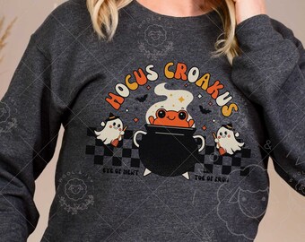 May include: Dark heather gray sweatshirt featuring a Halloween-themed graphic. The design includes a cauldron with a frog, ghosts, and the text "Hocus Croakus." The sweatshirt has a crew neck and long sleeves, perfect for the fall season.