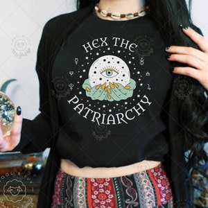 Puede incluir: Sudadera negra con el texto "Hex the Patriarchy" y un gráfico de una mano sosteniendo una bola de cristal con un ojo dentro. El gráfico está rodeado de estrellas y símbolos.