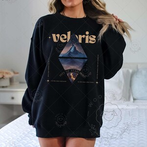 Puede incluir: Sudadera negra con un dise&ntilde;o gr&aacute;fico que presenta una cordillera y el texto "velaris city of starlight" en una fuente dorada.