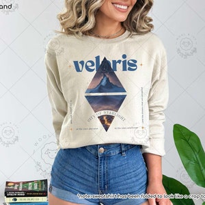Puede incluir: Una sudadera beige con un dise&ntilde;o gr&aacute;fico azul que presenta una cordillera y el texto "velaris - CITY OF STARLIGHT - at the stars and with the stars whellisten and the dreams that are answered to the people who look".