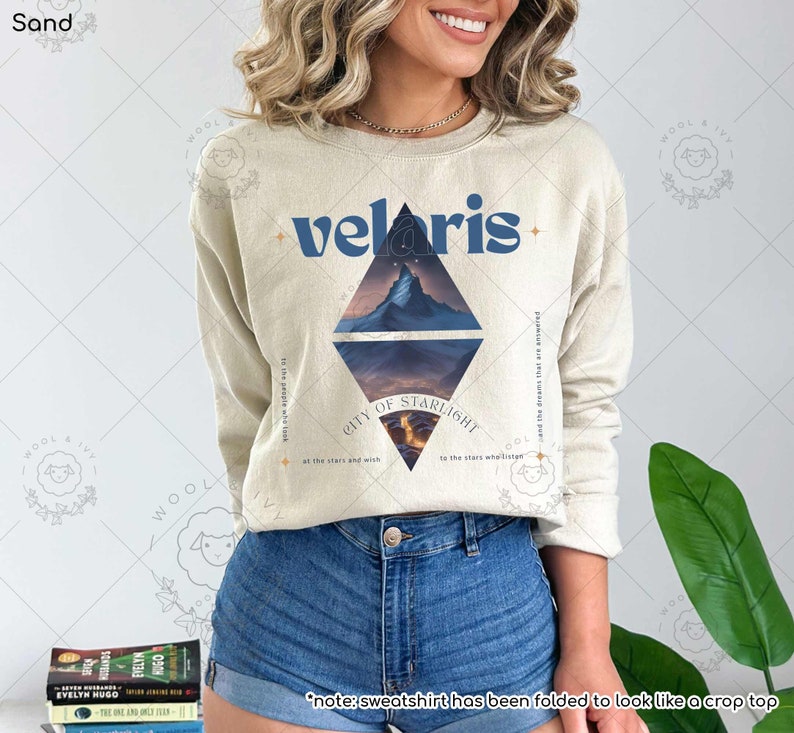 Puede incluir: Una sudadera beige con un dise&ntilde;o gr&aacute;fico azul que presenta una cordillera y el texto "velaris - CITY OF STARLIGHT - at the stars and with the stars whellisten and the dreams that are answered to the people who look".