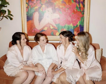 May include: Four women in matching light pink robes with lace trim sit on a rust-colored couch. A colorful painting hangs on the wall behind them. The women are smiling and appear to be enjoying each other's company.
