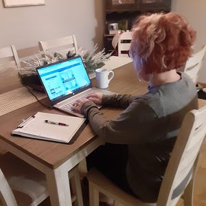 May include: A person is seated at a wooden table, working on a laptop. A white mug sits to the right of the laptop. A clipboard with a pen rests on the table. The person is wearing a long-sleeved shirt.