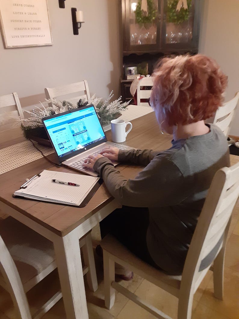May include: A person is seated at a wooden table, working on a laptop. A white mug sits to the right of the laptop. A clipboard with a pen rests on the table. The person is wearing a long-sleeved shirt.