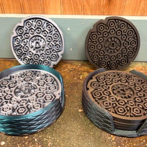 May include: Set of four coasters with a manhole cover design. Two coasters are silver, two are brown. Each coaster has a detailed geometric pattern and is held in a matching holder with a textured, braided design.