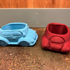 May include: Two car-shaped planters, one light blue and one red, sit side-by-side. The planters are designed to resemble classic Volkswagen Beetles, with rounded bodies, headlights, and wheel wells. The planters are made of a textured material.