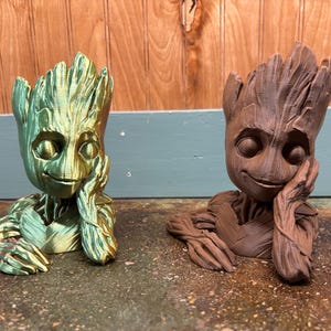 May include: Two Groot-inspired figurines, one in metallic green and the other in brown, are displayed. Each figure has a thoughtful expression, resting their head on a hand. The figures are likely decorative items or planters.