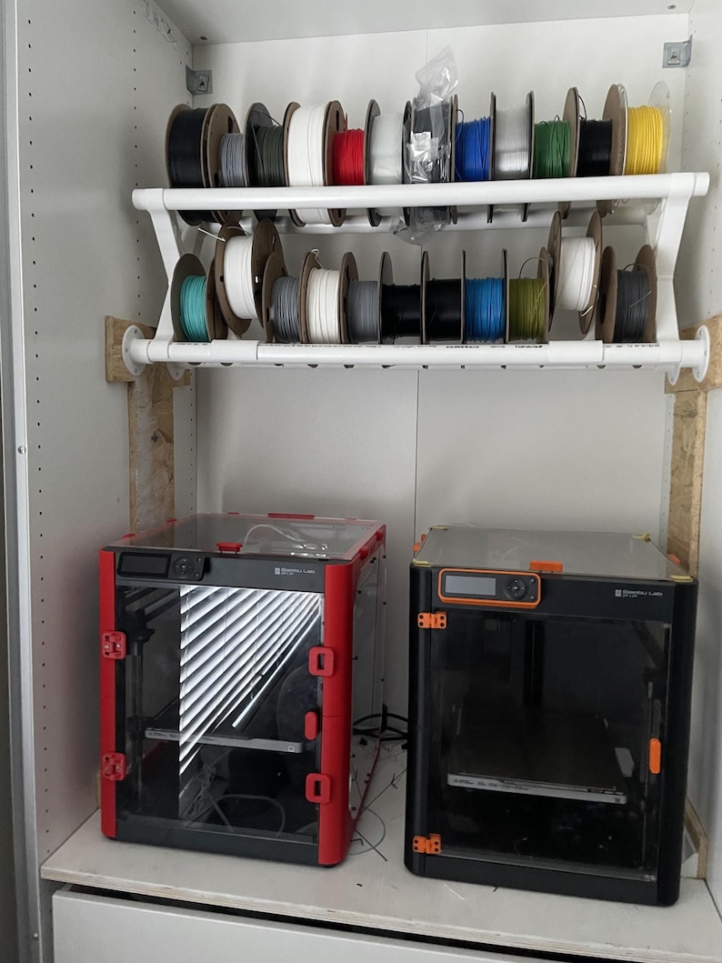 May include: Two 3D printers, one red and one black, are on a shelf in a cupboard. Above the printers is a shelf with spools of filament in various colours, including red, blue, green, and yellow.