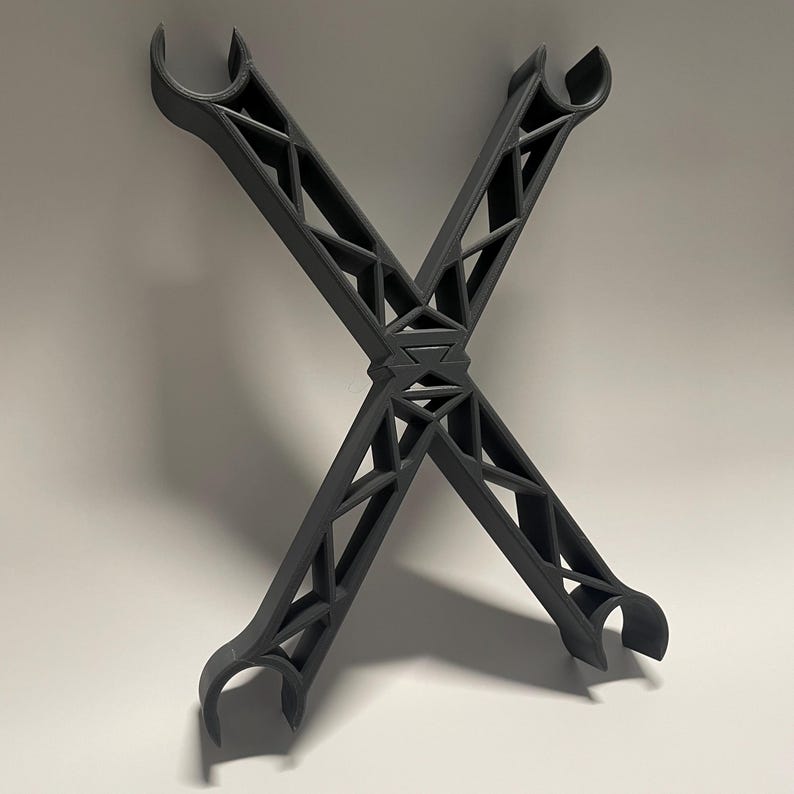May include: A black, 3D printed, X-shaped structure with a geometric design. The structure has two arms that are bent at a 90-degree angle, creating a hook-like shape at each end.