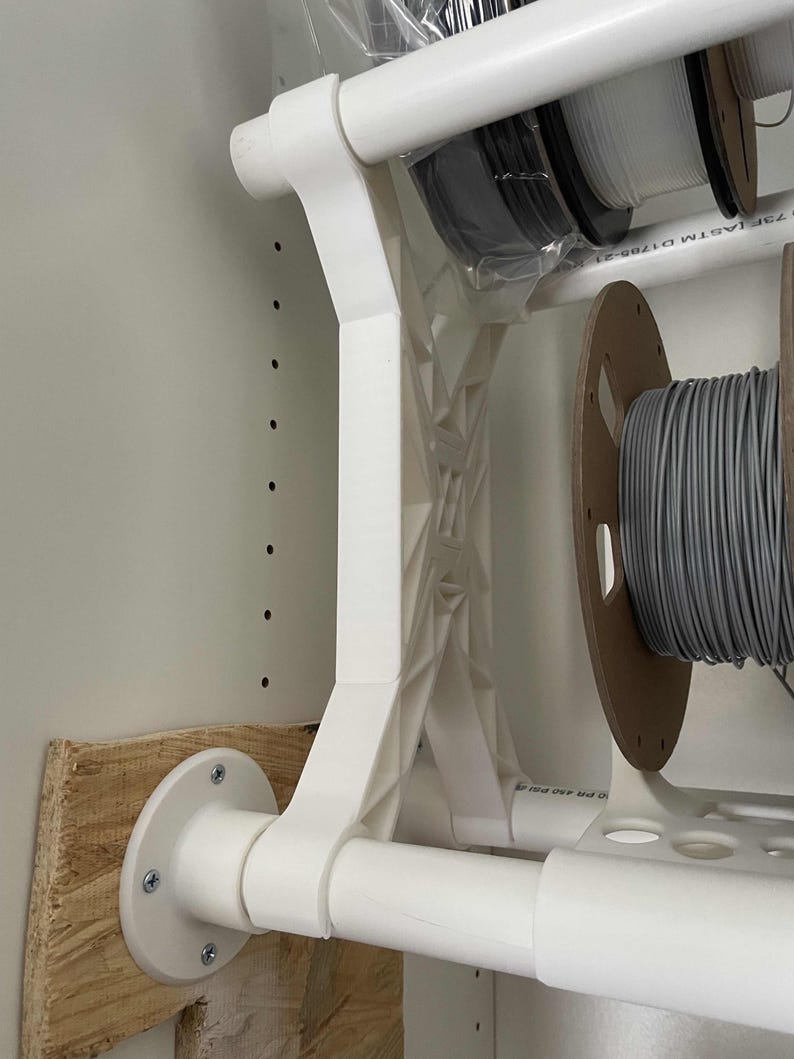 May include: White plastic shelf brackets with a white pipe attached to the bracket. The bracket is mounted to a wooden board with screws. The pipe is labelled "25F 1/2" and "ASTM D1985-21".