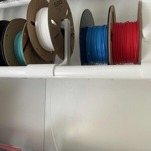 May include: White, blue and red filament spools are organised on a white shelf with a white plastic holder. The spools are labelled "Elegoo".