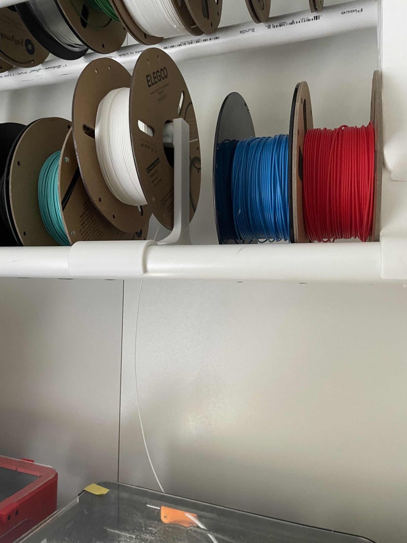 May include: White, blue and red filament spools are organised on a white shelf with a white plastic holder. The spools are labelled "Elegoo".