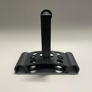 May include: Black plastic stand with a vertical post and a curved base with multiple holes. The stand has the letters "NM" printed on the front.