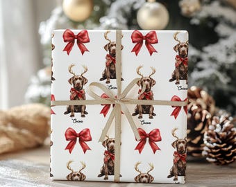 May include: Wrapped gift with white paper featuring a repeating pattern of brown dogs wearing antlers and red bows. The gift is tied with a gold ribbon and bow, perfect for Christmas or holiday gifting.