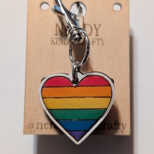May include: A heart-shaped keychain with a rainbow design, attached to a silver clasp. The heart features horizontal stripes in red, orange, yellow, green, blue, and purple. The keychain is on a brown tag with the text "Nerd Kind Crafty".