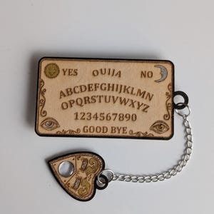 Peut inclure: Une planche Ouija en bois avec des lettres, des chiffres et des symboles gravés, dont des yeux, un soleil et une lune. La planchette en forme de cœur est reliée par une chaîne argentée. La planche indique "OUI", "NON" et "AU REVOIR".