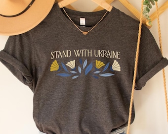 May include: A dark gray V-neck t-shirt with the text "Stand with Ukraine" and a floral design in blue, yellow, and white. The shirt is hanging on a wooden hanger and is styled with denim shorts and a straw bag.