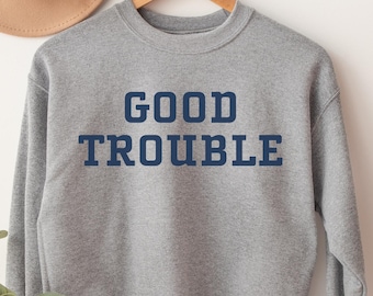 Puede incluir: Una sudadera gris con el texto "GOOD TROUBLE" impreso en azul marino.