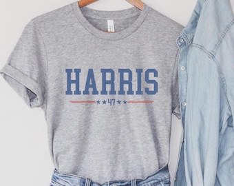 May include: Gray t-shirt with the text "HARRIS" in blue with red and white stars and the number "47" underneath. The shirt is paired with denim shorts and a light blue denim shirt.