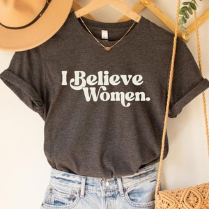 Puede incluir: Una camiseta gris oscuro con el texto "I Believe Women." impreso en blanco. La camiseta está colgada de una percha de madera y se combina con un sombrero marrón y un bolso de crochet.
