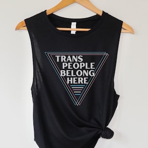Op de afbeelding: Zwart sleeveless muscle tank top met een witte driehoek grafische en de tekst "TRANS PEOPLE BELONG HERE" in een wit lettertype. De driehoek grafische is omlijnd in roze, blauw en lichtblauw.