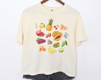 Op de afbeelding: Een crèmekleurig T-shirt met een afbeelding van verschillende vruchten, waaronder een ananas, papaja, druiven, aardbeien en kiwi. De tekst "FRUITY" is op het shirt gedrukt.