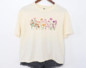 May include: Cream-colored t-shirt featuring a watercolor floral design across the chest. The flowers are in shades of pink, orange, yellow, and purple. The shirt is hanging on a wooden hanger against a white background. Black denim shorts are visible below the shirt.