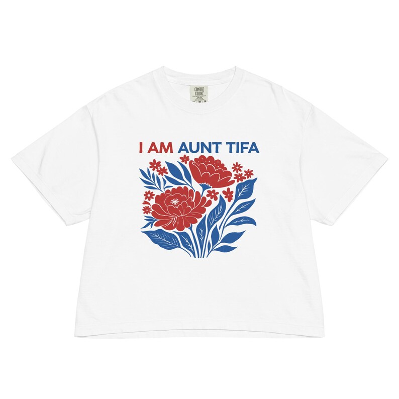 May include: White t-shirt with the text "I AM AUNT TIFA" in red and blue. The shirt features a floral design with red flowers, blue leaves, and small red star-shaped flowers. The shirt is made of a soft, comfortable material.