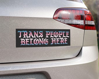 May include: A black bumper sticker with white text that reads "Trans People Belong Here" in a gothic font. The text is outlined in pink and blue.