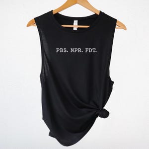Puede incluir: Camiseta sin mangas negra con la inscripción "PBS. NPR. FDT." en blanco. La camiseta está colgada de una percha de madera sobre un fondo blanco. La parte inferior de la camiseta está anudada.