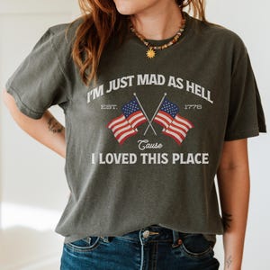 May include: Dark olive green t-shirt with the text "I'M JUST MAD AS HELL CAUSE I LOVED THIS PLACE" and two crossed American flags. The shirt includes the year 1776 and the word "EST."