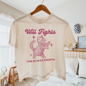Könnte beinhalten: Cremefarbenes T-Shirt mit einem rosa Aufdruck einer kämpfenden Katze, begleitet vom Text "Will Fights FOR HUMAN RIGHTS". Das Design enthält Sterne. Das Shirt hängt an einem Holzbügel.