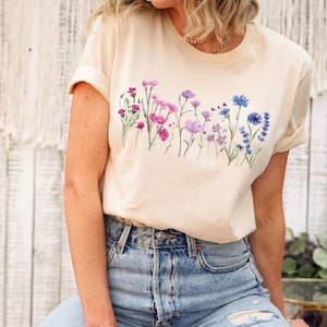 Può includere: Una t-shirt color crema con un design floreale con fiori rosa, viola e blu.