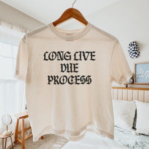 Puede incluir: Camiseta color crema con la frase "LONG LIVE DUE PROCESS" impresa en letras g&oacute;ticas negras. La camiseta est&aacute; colgada en una percha de madera sobre un fondo neutro. Es una prenda informal.
